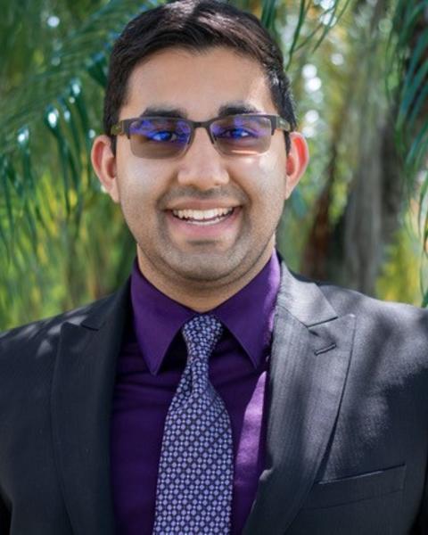 Headshot of Daniel in a dark suite with a purple shirt and tie, standing in front of a palm tree backgroun.