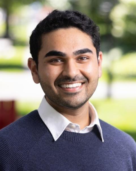 Headshot of Ishan in a navy sweater with a blurred outdoor background facing the viewer