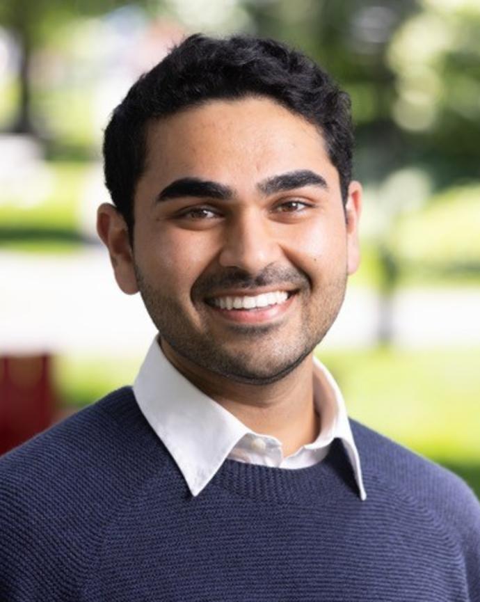 Headshot of Ishan in a navy sweater with a blurred outdoor background facing the viewer